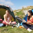Three women smile while eating lunch in front of camping tents on a sunny day
