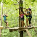 A mother and two daughters stand on wooden platform and watch dad navigate a treetop obstacle