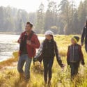 Smiling family of four wearing hiking gear walk along sunny lakeshore in a forest