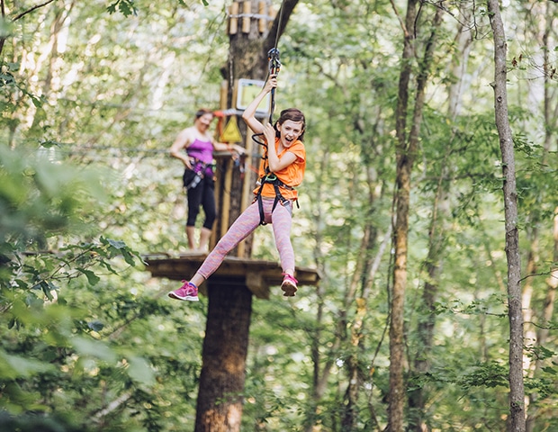 Mom watches from wooden platform while daughter rides zipline, smiling and gripping her safety lines
