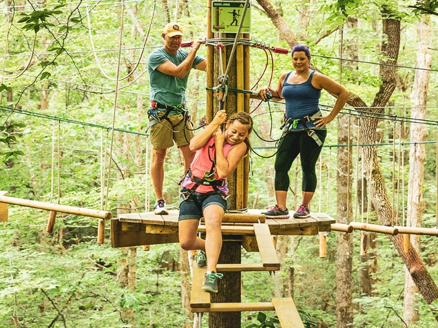 Young girl intently navigates treetop obstacle while parents watch from adjacent wooden platform