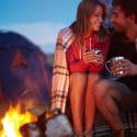 Affectionate share a blanket and couple clasp mugs of hot cocoa in front of a campfire and tent while the sun sets