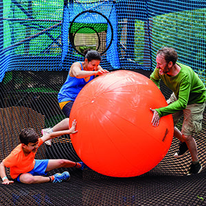 A man, woman, and child stand on suspension nets and playfully wrestle a large rubber ball from each other's grasp