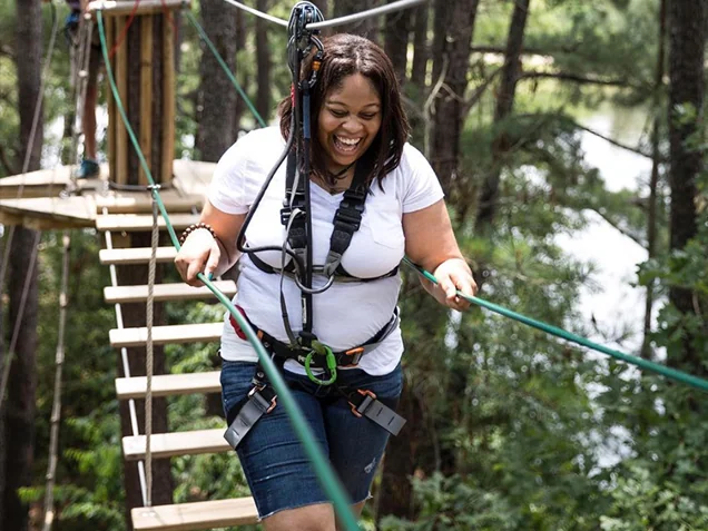 Young woman grins while climbing across a treetop bridge