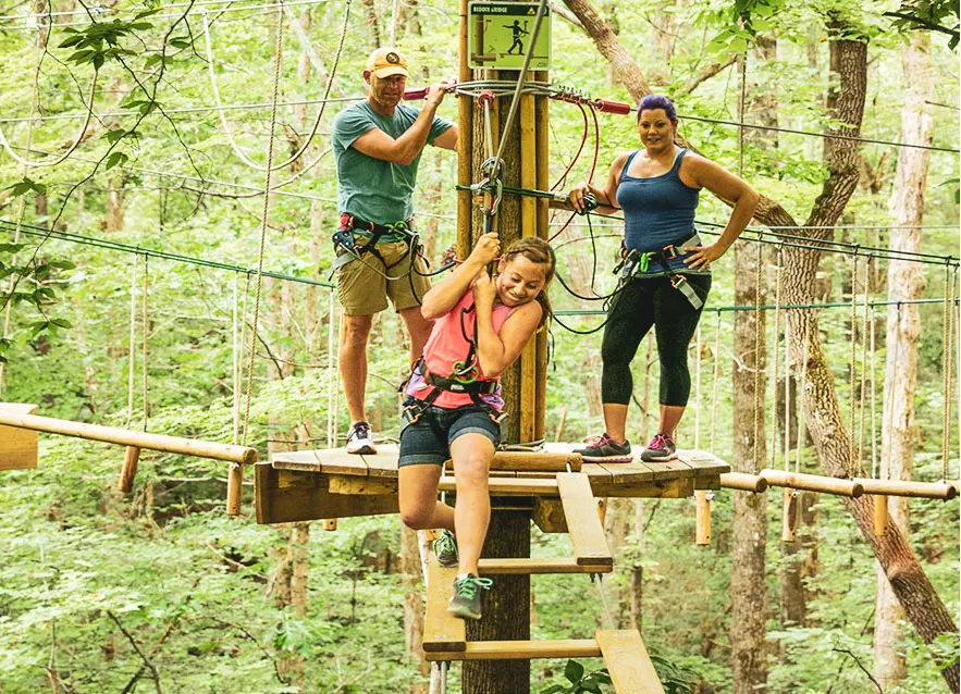 Young girl intently navigates treetop obstacle while parents watch from adjacent wooden platform