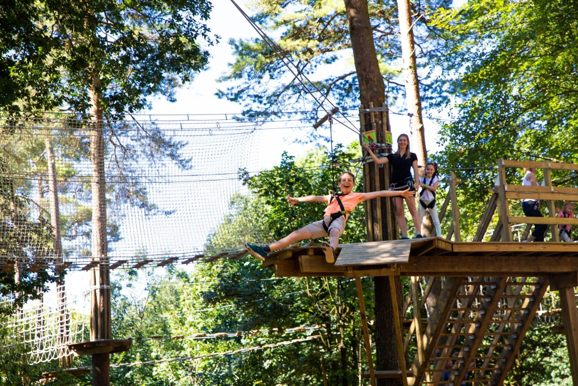 Young boy stretches out his arms as he flies down a Go Ape Treetop Adventure zipline with his Mom and sister watching from the treetop platform