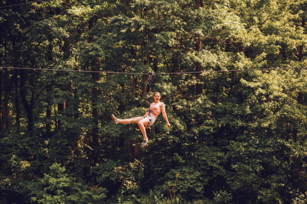 Girl smiles at the camera while descending a zipline in front of a dense forest