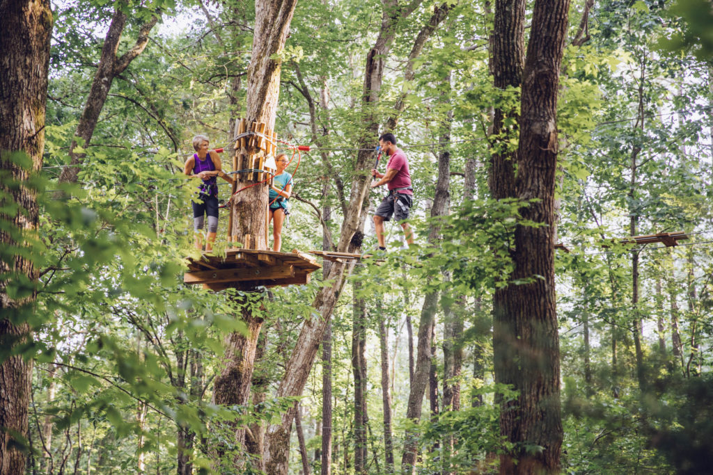 Two women stand on a wooden platform and watch a man navigate a suspended obstacle