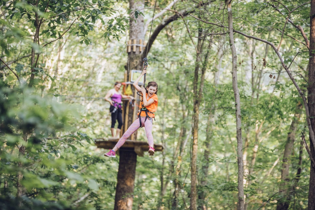 Young girl holds her safety lines and yells excitedly while riding a zipline away from a woman standing on a wooden platform