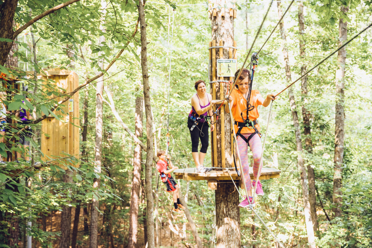 Woman on wooden platform laughs while watching a young girl balance on a single rope while traversing a treetop obstacle