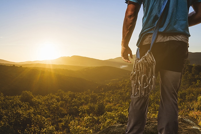 Man dressed in rock climbing gear watches the sun rise over a forest valley