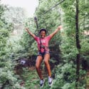 Young woman excitedly throws her arms up in the air while riding zip line through a forest