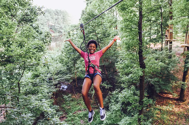 Young woman excitedly throws her arms up in the air while riding zip line through a forest