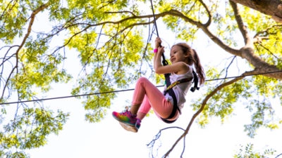 Small girl raises her legs and clutches her safety lines and glides through a treetop obstacle instead of walking through